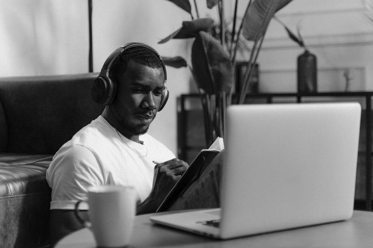 A Grayscale of a Man Writing on His Planner while Wearing Wireless Headphones